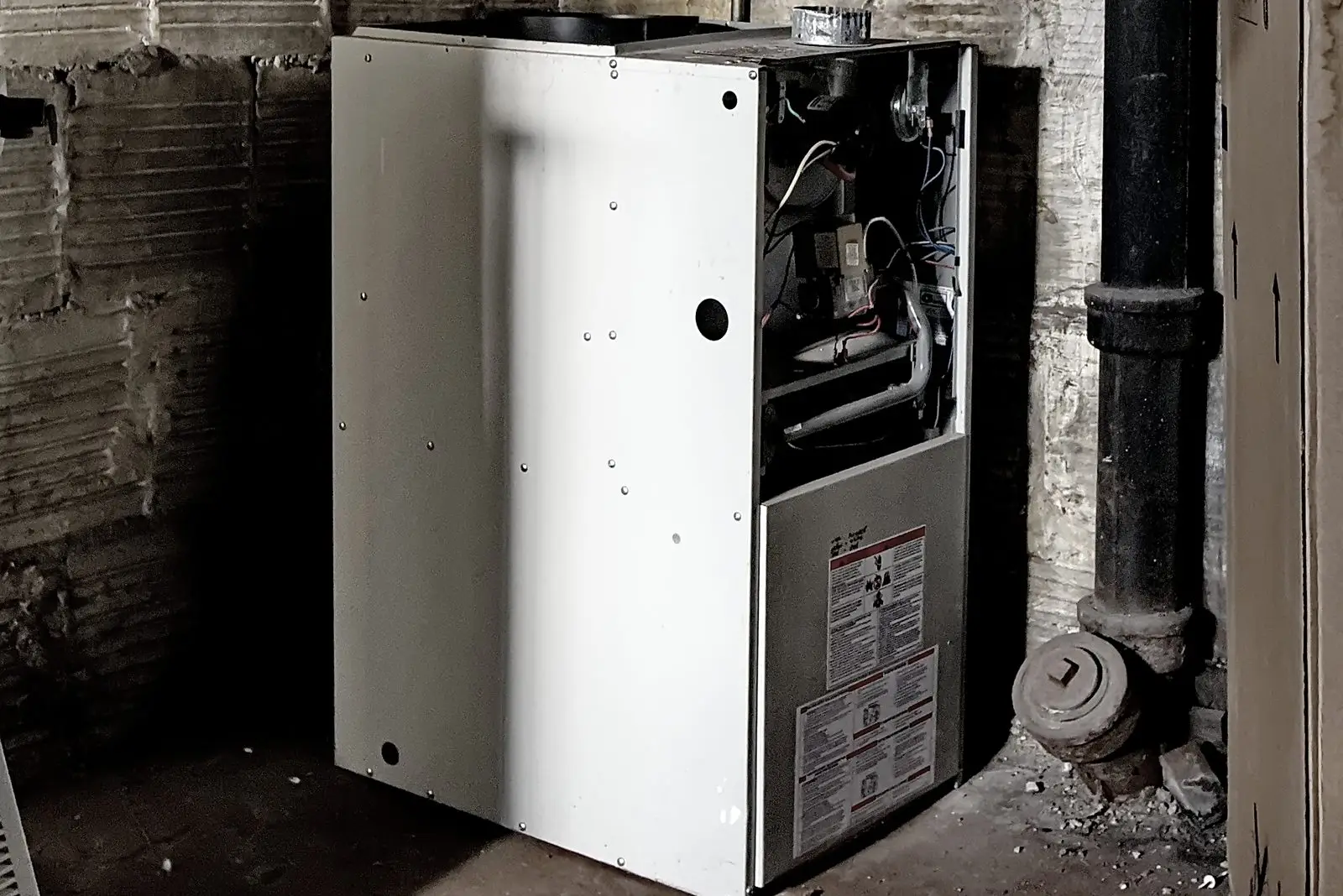 Gas furnace with its upper panel off in an old brick basement beside a cast iron pipe