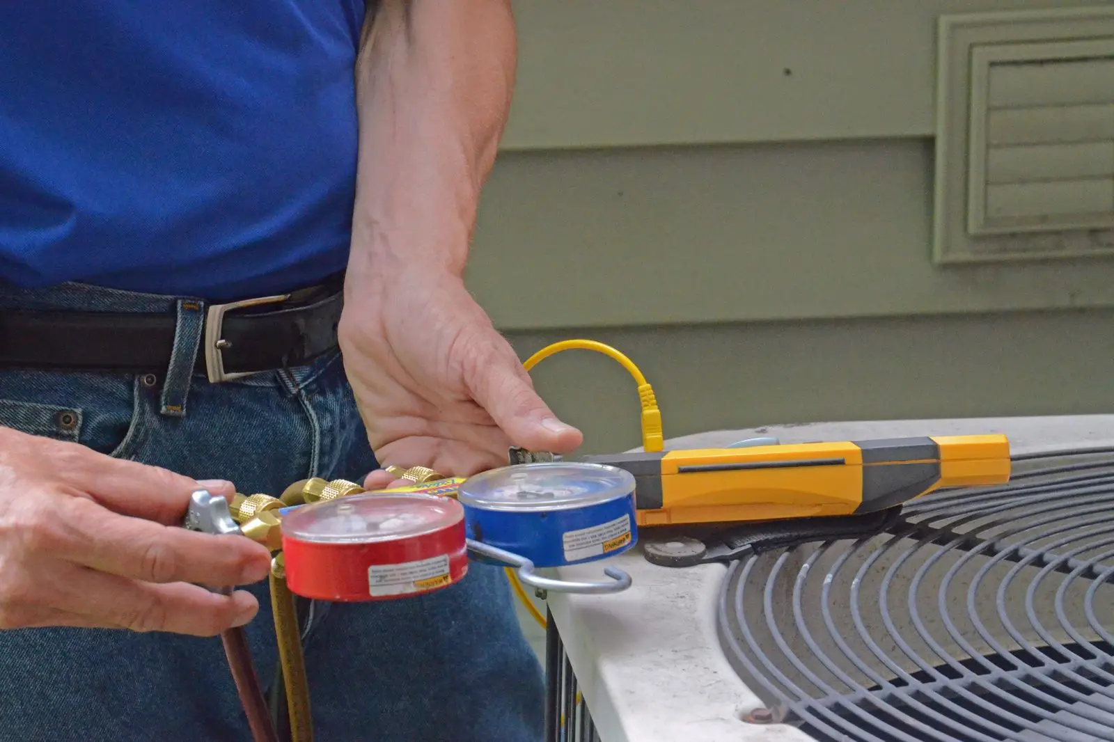 Technician holding refrigerant gauges beside an outdoor AC condenser during a tune-up